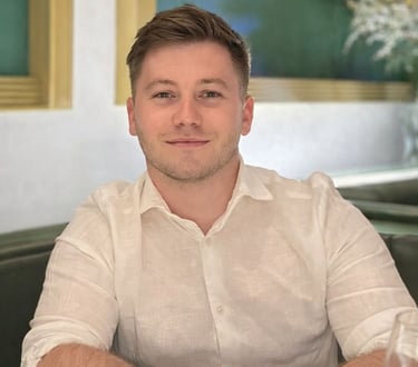 Harrison, smiling, wearing a white linen shirt and silver watch sitting at a restaurant table.