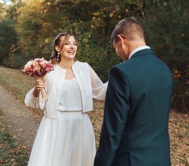 Le marié découvre la mariée et lui tend la main, tous deux souriants au cœur d'une allée automnale.