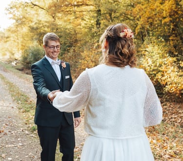 Le marié découvre la mariée et lui tend la main, tous deux souriants au cœur d'une allée automnale.