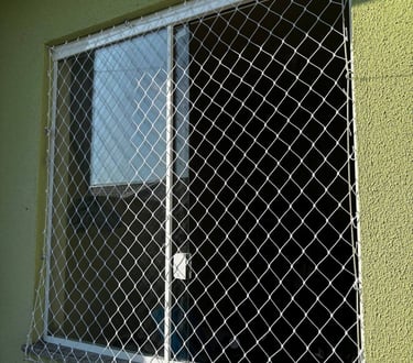 janela de correr de alumínio em parede verde com rede de proteção instalada externamente; luz do sol incidindo e fixações vis