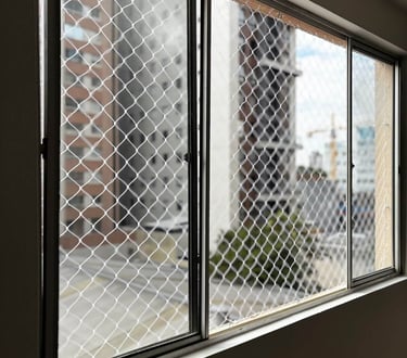 janela de correr de alumínio vista por dentro com rede de proteção instalada; ao fundo, prédios residenciais em dia claro