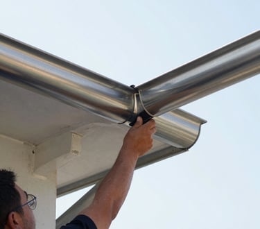Close-up of a worker installing a clean stainless steel gutter on a high roofline, bright natural outdoor daylight, sharp focus on the metallic reflection, 35mm lens