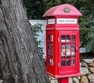 Red British phone-booth style Book Exchange box filled with books next to a large tree trunk.