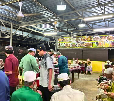 Muslim community gathering at an outdoor food stall for a communal Halal dinner event under a metal roof.