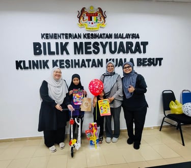Staff and children pose at Klinik Kesihatan Kuala Besut with a toy scooter and health campaign posters.