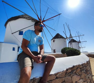 A man in sunglasses sitting before iconic Mykonos windmills under a bright sunny sky in Greece.