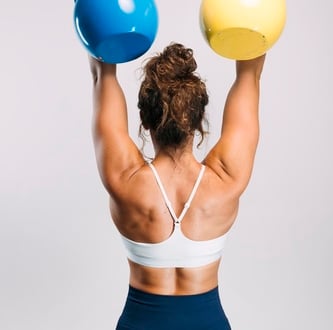 Athletic woman lifting blue and yellow kettlebells overhead for a strength training back workout.