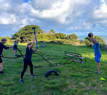 People performing outdoor functional fitness training with battle ropes and slam balls on a coastal lawn.