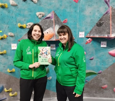 two women in green jackets standing next to a climbing wall and holding a book