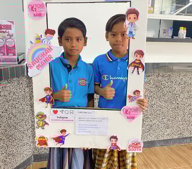 Two young boys in blue shirts giving thumbs up inside an Instagram-themed photo booth frame at Klinik Raudhah Manir.