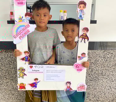 Two young boys posing in a decorated photo booth frame at Klinik Raudhah Manir healthcare center.