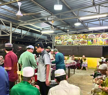 Muslim community gathering at an outdoor food stall for a communal Halal dinner event under a metal roof.