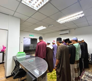 A group of Muslim men performing congregational prayer in an office prayer room on green prayer mats.
