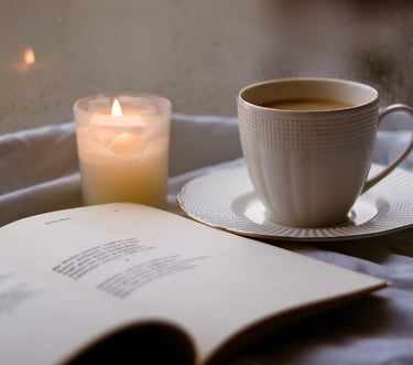 A cozy scene featuring an open book, a cup of hot coffee, and a lit candle on a bed.