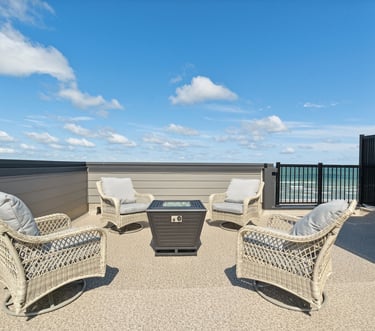 Wicker patio chairs surrounding a modern fire pit on a rooftop deck with beachfront views.