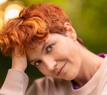 Portrait of a person with curly red hair and hazel eyes posing outdoors with soft bokeh lighting.