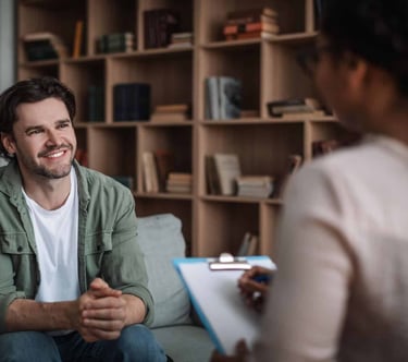 A smiling man sits on a sofa during a mental health therapy session with a counselor.