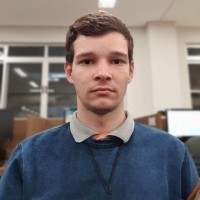 Pessoa usando suéter azul com gola de camisa cinza, em ambiente interno com janelas e computadores.