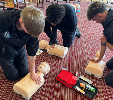3 people kneel on the floor practicing CPR on adult training manikins, with an AED