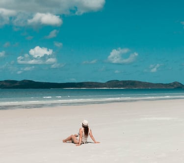 A woman wearing a sun hat sits on a white sand tropical beach with blue ocean water and a cloudy sky.