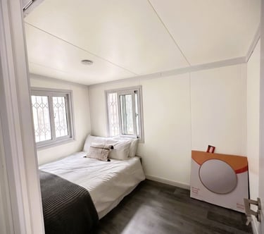 A minimalist tiny house bedroom with a white bed, dark wood floors, and dual windows.