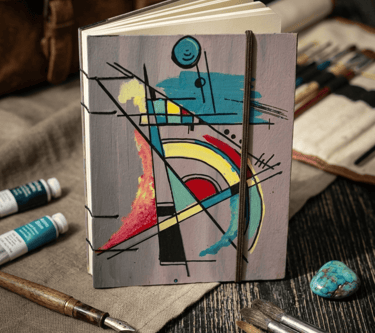 Cuaderno de arte abstracto pintado a mano rodeado de pinceles, tubos de acrílico y plumas