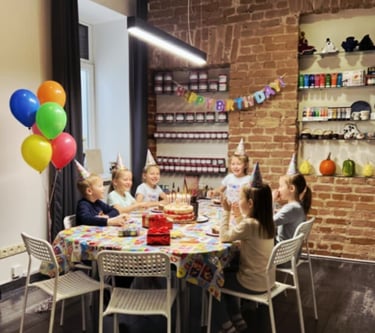 Group of happy children wearing party hats celebrating a birthday around a table with cake and balloons.