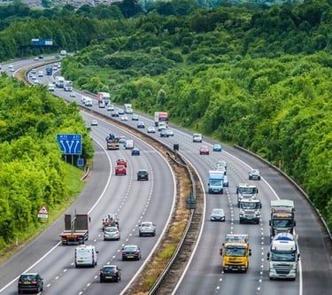 Busy traffic with cars and trucks driving on the M25 motorway through a lush green landscape.