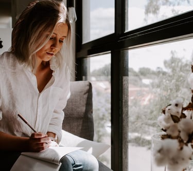 A woman in a white linen shirt writing in a journal by a bright window with cotton stems.