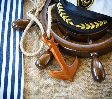 A decorative captain's hat, placed on a ship's wheel with an ornamental wooden anchor next to it.