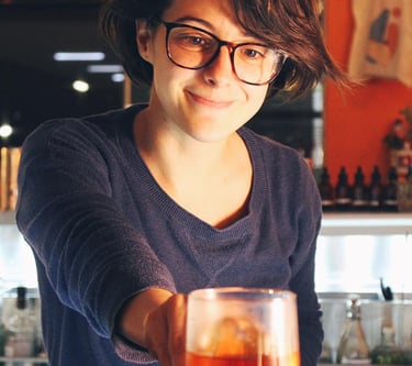 Smiling female bartender serving a craft cocktail in a glass at a dimly lit bar counter.