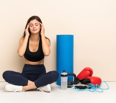 A woman in fitness wear sitting with closed eyes next to a yoga mat, boxing gloves, and workout gear.
