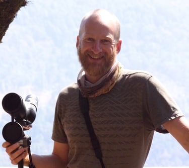 San Cristobal bird guide Benedict Simmons smiling with telescope, with binoculars in front hillside