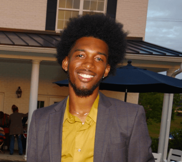 Kelvin in a suit and yellow shirt standing in front of a house