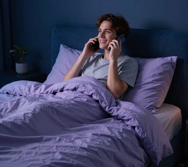 A serene North American bedroom at night. A person looks relaxed and happy while talking on a phone, wrapped in a soft blanket. The colors are deep midnight blue and soft lavender, suggesting premium comfort and support.