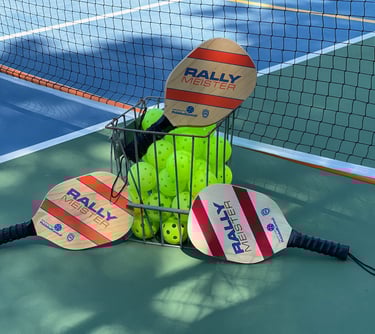 Three Rally Meister wooden pickleball paddles leaning against a metal basket filled with neon yellow pickleballs on a court.