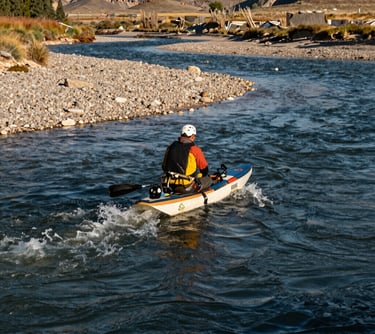 An outdoor action shot of a guide on a Montana river. The water is a deep slate blue, the banks are earthy off-white gravel, and the lighting is the bright, clear gold of a Montana morning.