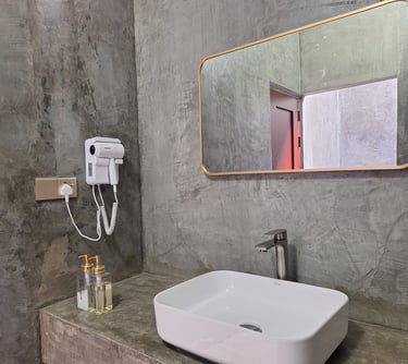 Modern industrial bathroom with a white vessel sink, gold framed mirror, and hair dryer against concrete walls.