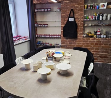 Pottery studio workshop table with unpainted ceramic clay pieces, brushes, and paints against a rustic brick wall.
