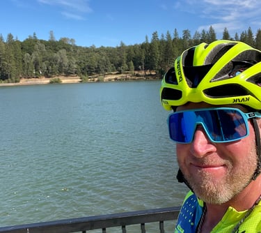 A picture of Justin in cycling gear standing next to a body of water (lake).