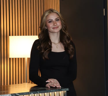 Smiling young woman with wavy blonde hair in a black dress posing by a marble counter.