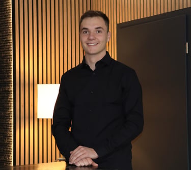 Smiling young businessman in a black button-down shirt posing in a modern office with wood paneling.