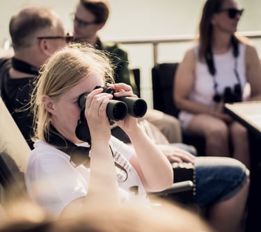 A blonde woman uses black binoculars to look at a distance during a sunny boat tour with group.