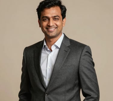A professional corporate headshot of a South Asian man in his late 30s, wearing a tailored charcoal suit, smiling confidently, wealth consultant style, warm studio lighting.