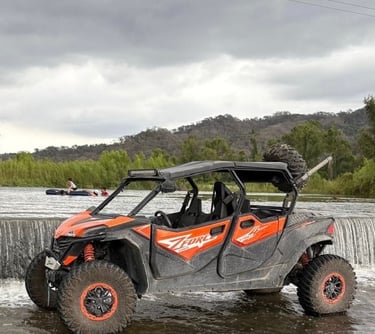 Orange CFMOTO ZFORCE side-by-side UTV parked in a shallow river by a waterfall under cloudy skies.