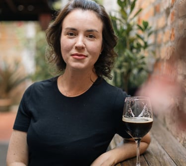A woman in a black shirt sits at a rustic outdoor bar with a glass of dark craft beer.