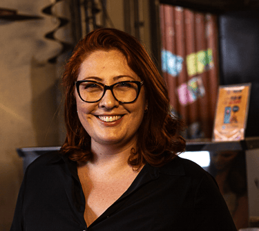 Smiling woman with red hair and black glasses wearing a dark button-down shirt in a cafe.