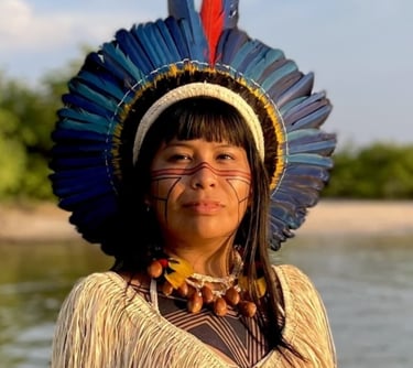 Indigenous Brazilian woman wearing a blue feather headdress and traditional facial paint by a river.