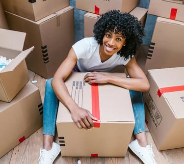 Smiling woman with curly hair hugging a cardboard moving box in her new home.