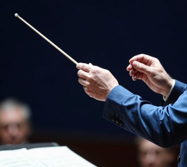 A portrait of a conductor's hand holding a baton, captured mid-motion during a rehearsal. The style is professional and focused, with a blurred dark navy blue background and sharp steel blue highlights on the sleeve.